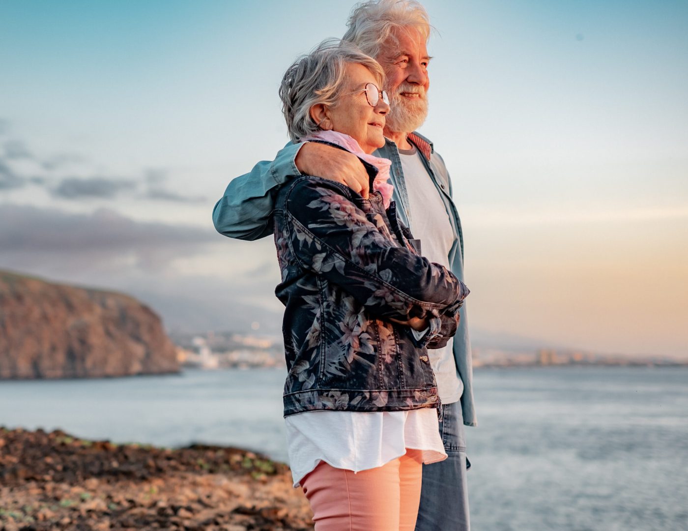 Lovely senior couple standing on the rocks at sea enjoying nature and freedom looking the horizon at sunset light. Relaxed lifestyle for a caucasian couple of retirees